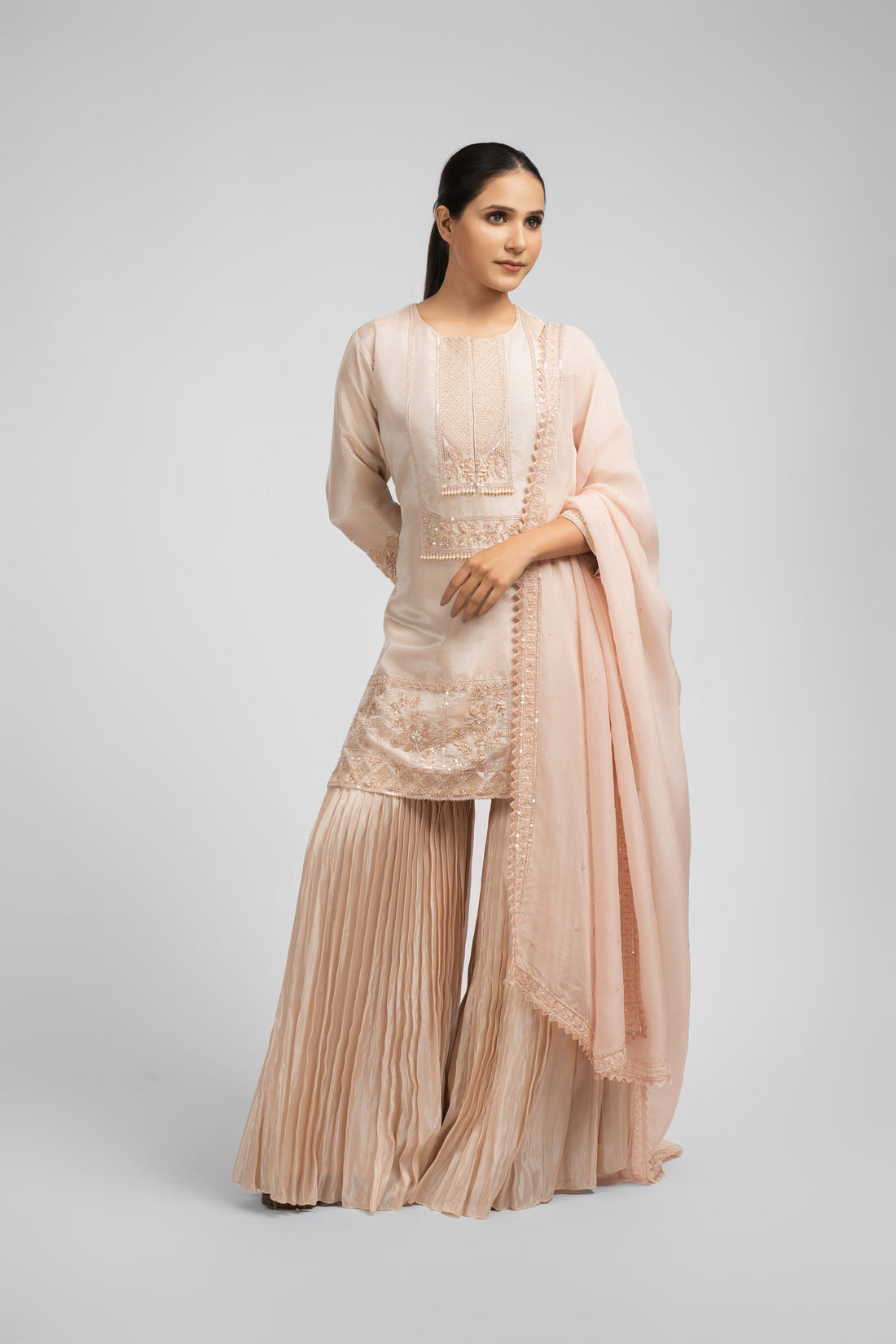 Model wearing an Ash Rose Pink tissue sharara suit with intricate embellishments on the kurta, paired with pleated sharara pants and an organza dupatta featuring pearl and sequin detailing, perfect for festive and formal wear.