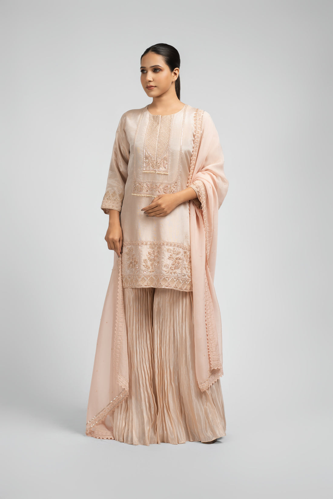 Model wearing an Ash Rose Pink tissue sharara suit with intricate embellishments on the kurta, paired with pleated sharara pants and an organza dupatta featuring pearl and sequin detailing, perfect for festive and formal wear.