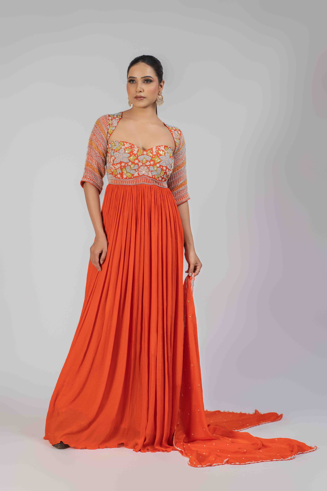 Orange chinnon anarkali gown with an embellished bodice, elbow-length sleeves, and an organza dupatta adorned with pearls, kasab, sequins, and colorful patchwork, ideal for festive and wedding occasions.