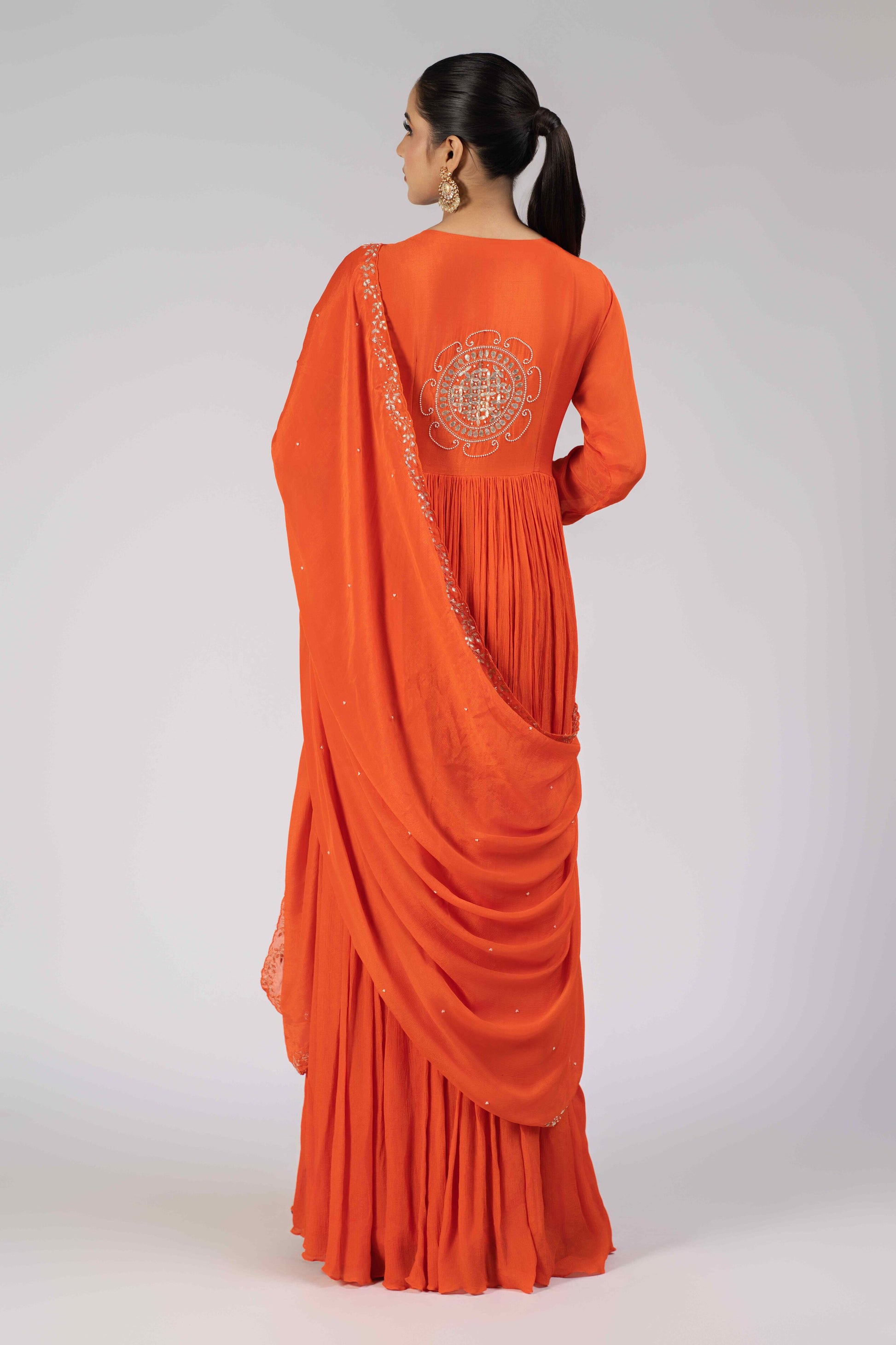 A model wearing an orange chinnon gota work gathered suit with an embellished bodice, sleeve cuffs, and a matching chinnon dupatta adorned with kasab, gotta patti, pearls, and sequins, perfect for festive and wedding occasions.