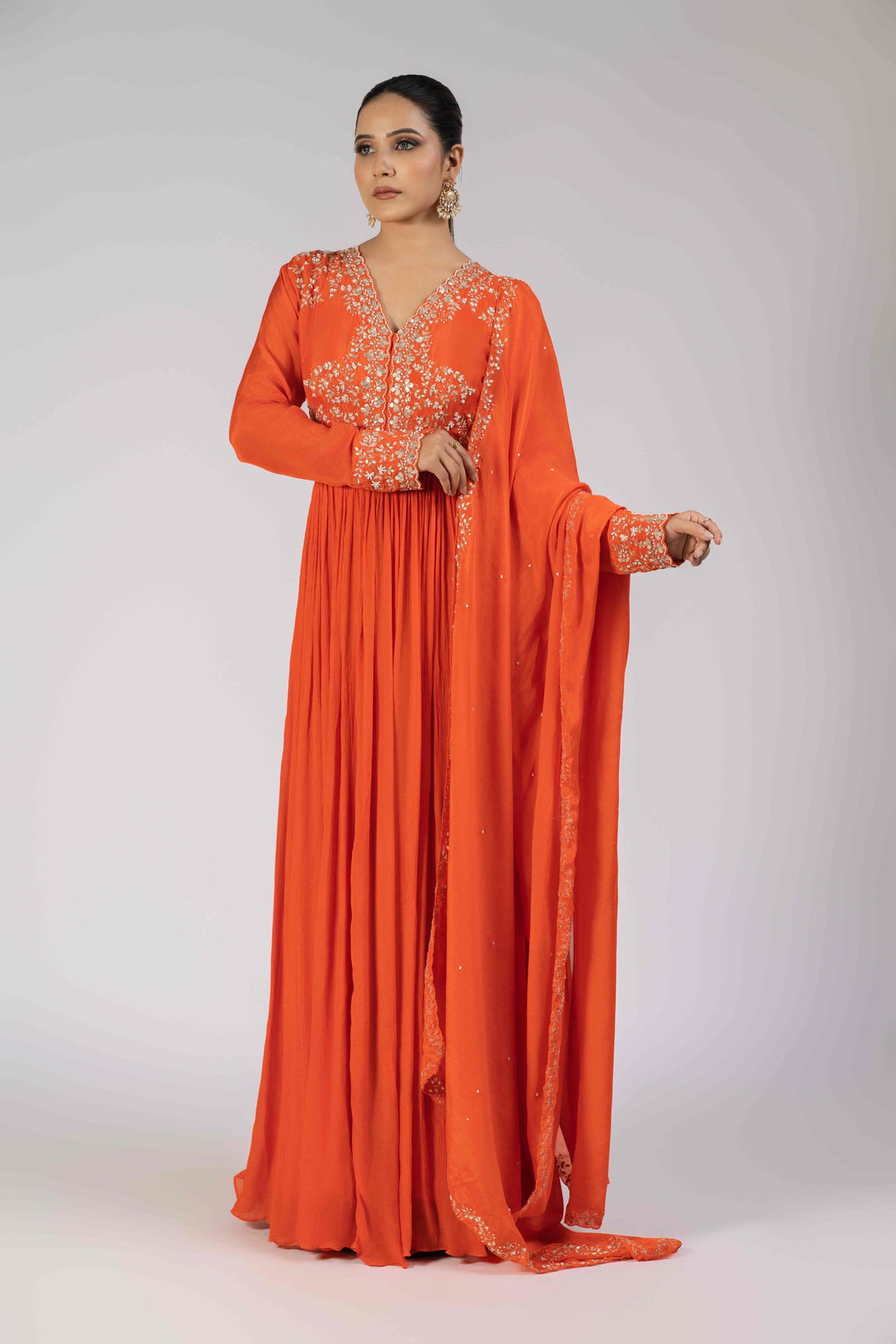 A model wearing an orange chinnon gota work gathered suit with an embellished bodice, sleeve cuffs, and a matching chinnon dupatta adorned with kasab, gotta patti, pearls, and sequins, perfect for festive and wedding occasions.