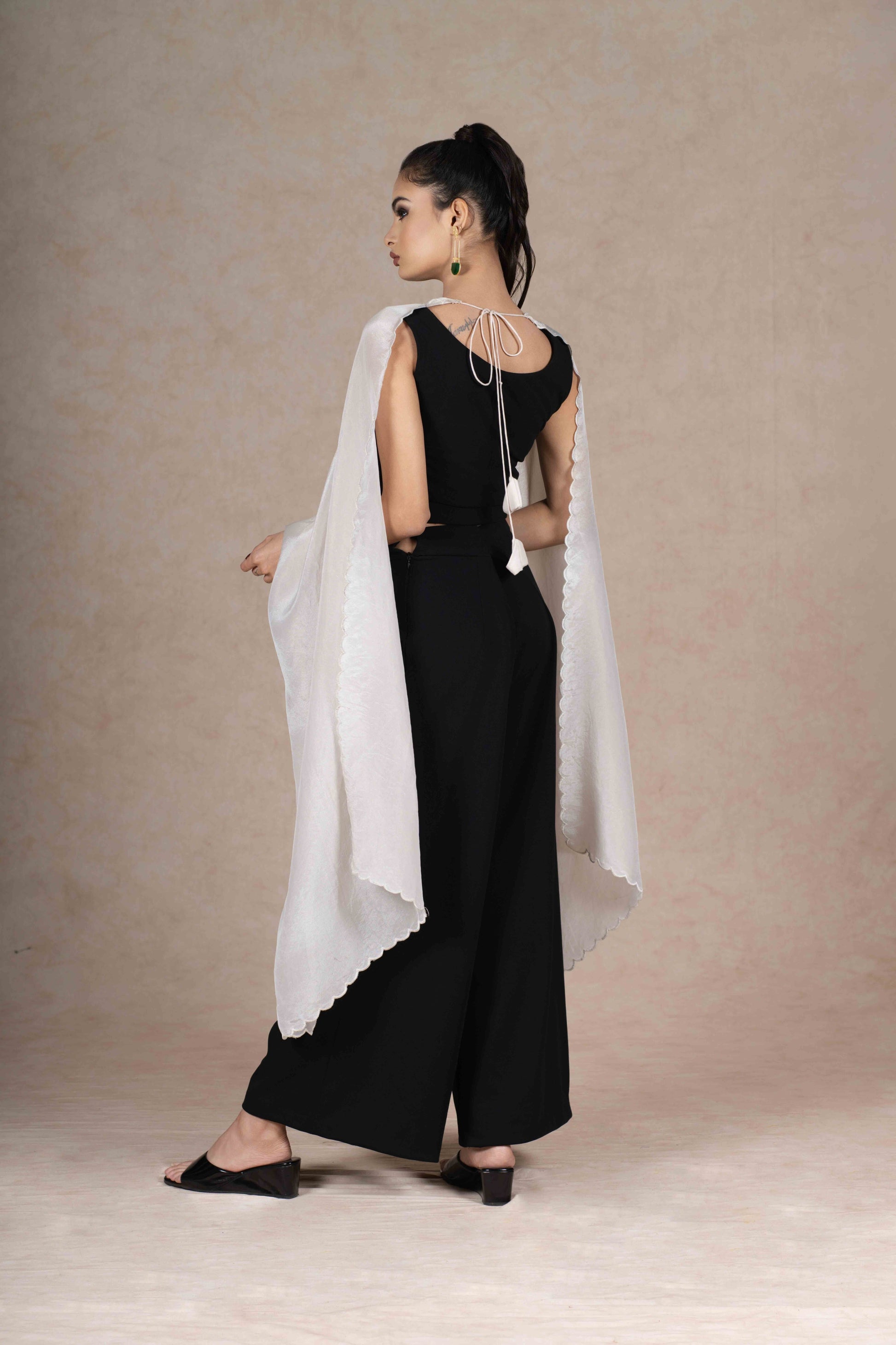 The flowing tissue cape extends gracefully from the back, highlighting its scalloped neckline detailing and hand-beaded embroidery, paired with smooth, slip-on black flared trousers.