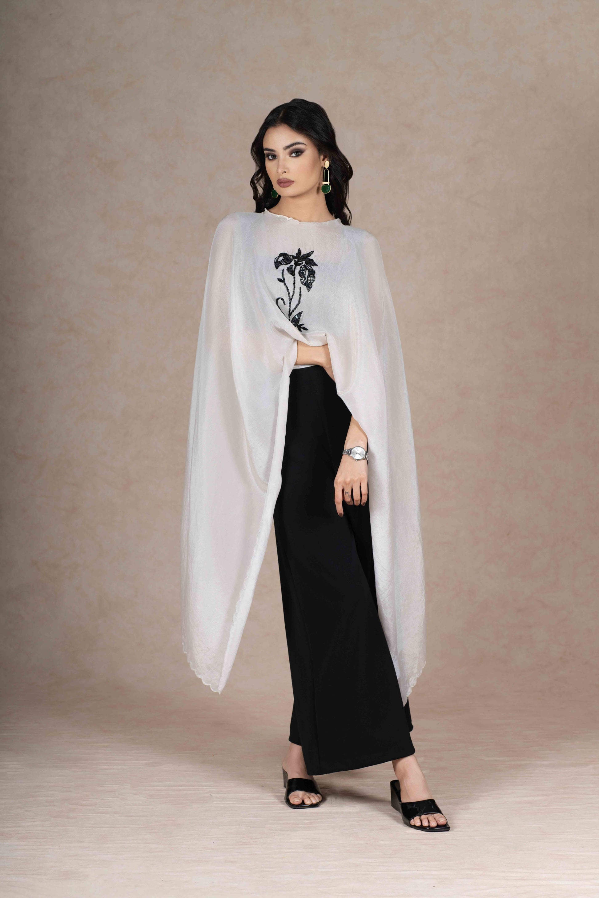 A sophisticated ensemble featuring a soft white tissue cape intricately hand-embroidered with black sequins, attached to black flared palazzo pants for a seamless, modern silhouette.