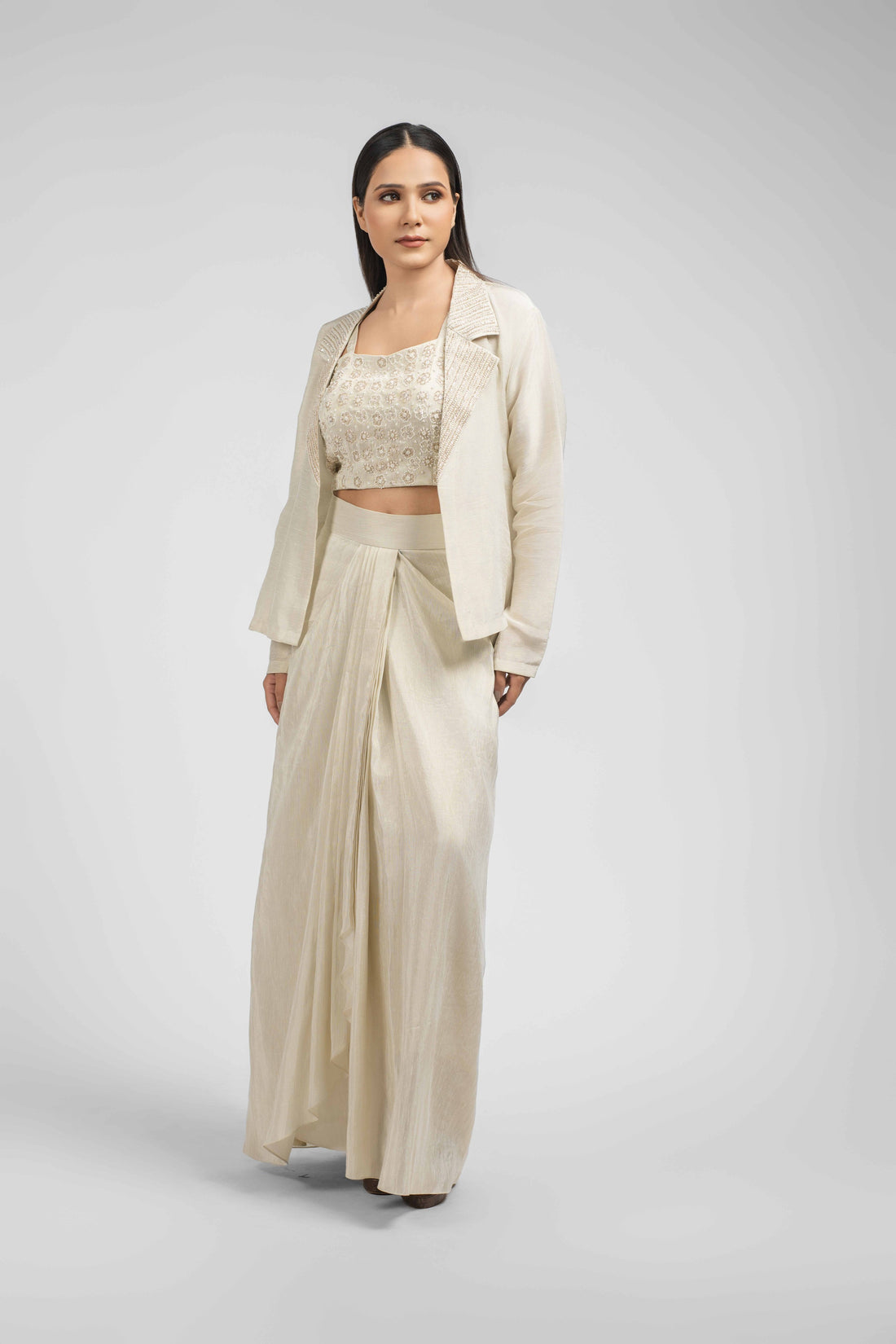 Ivory Linen Tissue Blazer Set with Drape Skirt featuring an embellished blazer, a sleeveless crop top, and a pleated dhoti skirt. Embellished with pearls, kasab, and cutdana, perfect for festive or formal occasions.