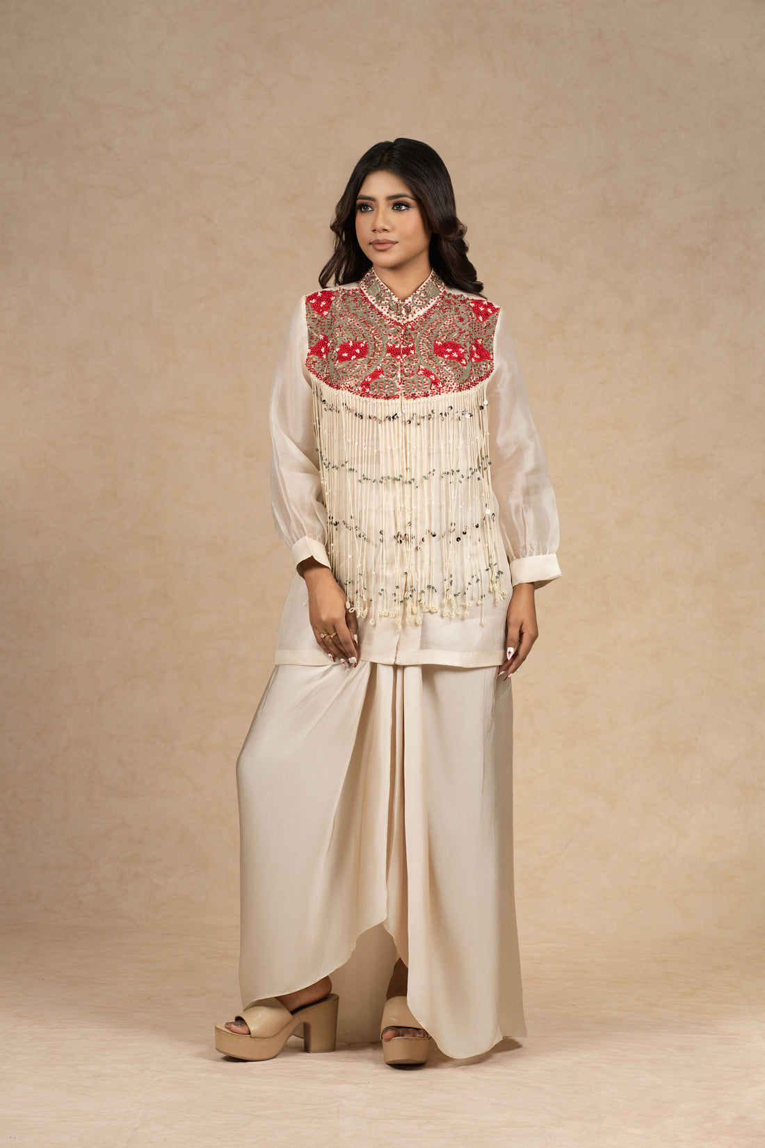  Model wearing an ivory organza Indowestern ensemble featuring a bustier and draped skirt, adorned with intricate dori work, pearl embellishments, and vibrant red embroidered detailing on the yoke.
