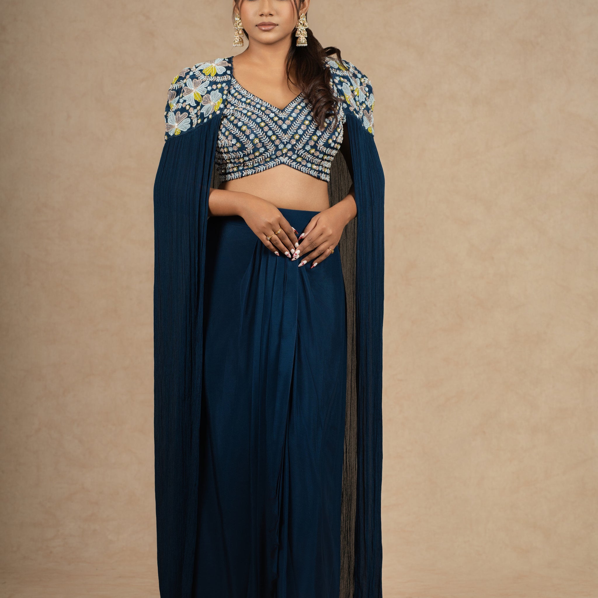 Model gracefully posing in a Royal Blue Indo-Western outfit featuring a heavily embellished blouse, pleated skirt, and a dramatic cape-style drape.