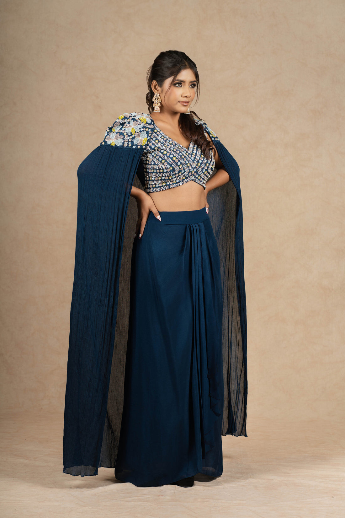 Model gracefully posing in a Royal Blue Indo-Western outfit featuring a heavily embellished blouse, pleated skirt, and a dramatic cape-style drape.